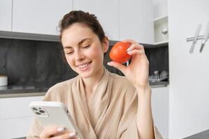 Image of smiling beautiful woman, sits in kitchen, holds smartphone and tomato, looks happy, orders groceries delivery to her home, using mobile phone app, buying online vegetables for her recipe photo