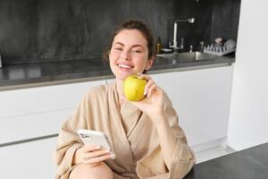 Close up portrait of happy young woman in bathrobe, sitting in the kitchen and using mobile phone, holding an apple, order fruits and vegetables online, using smartphone app for groceries delivery photo