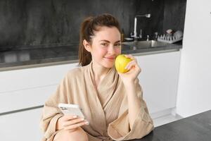 Image of attractive young woman in bathrobe, sits in kitchen, eats green apple and looks at mobile phone, uses smartphone app, orders food on application photo