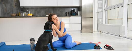 Portrait of beautiful brunette fitness girl, plays with her dog during workout training session, sits at home on rubber yoga mat, does sport exercises in living room photo