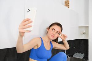 Portrait of smiling, beautiful fitness instructor, posing for fitness blog, selfie on smartphone, sitting in kitchen, looking happy photo
