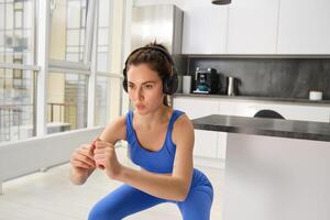 Image of young sportswoman, fitness instuctor doing squats workout at home, listening music in wireless headphones and stretching arms, doing exercises in living room photo