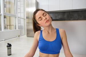 Portrait of young sportswoman, stretching her neck, warm-up before yoga exercises, doing fitness workout on rubber mat, wearing blue sportsbra and leggings photo