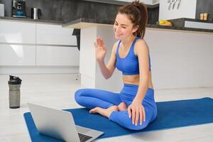 Portrait of fitness instructor, woman connects to online training session, waves hand at laptop, teaching yoga workout from home, sitting on rubber mat photo