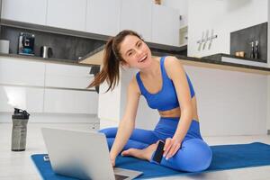 Portrait of young fitness girl paying for online class subscription, workout app, holding credit card, sitting on yoga mat with laptop, buying fitness tutorials photo