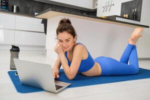 Portrait of young fitness woman, laying on rubber mat, using laptop, looking for workout video tutorials online photo