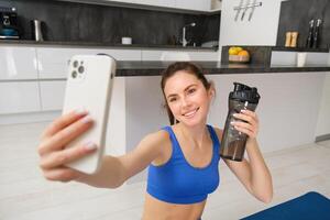 Wellbeing and sports. Beautiful young woman taking selfie on smartphone, doing fitness training from home, drink water, stays hydrated and doing exercises photo