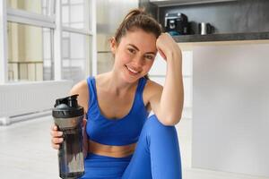 sonriente aptitud niña bebidas agua en el medio de formación sesión, sonriente y mirando feliz, rutina de ejercicio desde hogar en azul sujetador deportivo foto