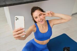 Portrait of young styling fitness girl doing workout from home, taking selfie and video for social media, gym instructor records her training session indoors photo