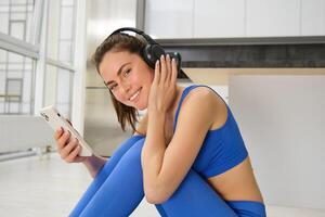 Fitness and wellbeing. Portrait of young woman doing fitness exercises, workout from home, wearing headphones and using smartphone, exercising indoors photo