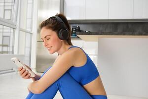 Portrait of brunette girl in fitness clothes, listens music in wireless headphones, holds smartphone, workout from home, looking at training video instructions photo