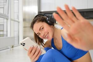 retrato de hermosa morena chica, se esconde desde cámara, pone su mano hacia cámara, escucha música en auriculares y sostiene teléfono inteligente, relojes ejercicios en línea en móvil teléfono, sonrisas foto