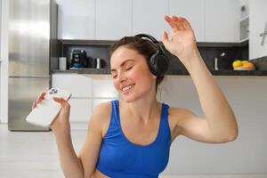 retrato de aptitud instructor, mujer con auriculares y teléfono inteligente, bailando a hogar, rutina de ejercicio dentro su casa, vistiendo azul sujetador deportivo foto