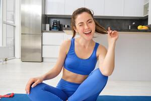 Image of young sport woman sitting at home on yoga mat, doing workout, stretching fitness exercises on floor in living room, smiling and looking happy at camera photo