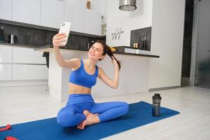 Sport and lifestyle concept. Young woman doing workout at home on yoga mat, makes fitness vlog with smartphone, takes selfie on mobile phone photo