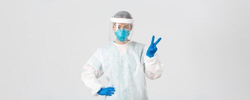 Covid-19, coronavirus disease, healthcare workers concept. Sassy asian female doctor, physician or tech lab in personal protective equipment showing peace sign, standing white background photo