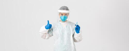 Covid-19, coronavirus disease, healthcare workers concept. Confident serious female asian doctor in personal protective equipment, show thumb-up and hold syringe with vaccine, white background photo