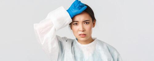Covid-19, coronavirus disease, healthcare workers concept. Young tired asian female doctor take-off personal protective equipment and wipe sweat, looking exhausted after long shift in hospital photo