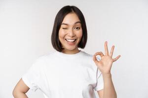 Everything okay. Smiling young asian woman assuring, showing ok sign with satisfied face, standing over white background photo