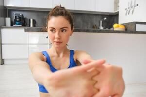 Portrait of beautiful woman does fitness training, sits on floor near kitchen, stretching her hands, workout from home photo