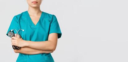 Covid-19, coronavirus disease, healthcare workers concept. Close-up of confident female asian physician, doctor in medical scrubs, cross arms determined and holding stethoscope, white background photo