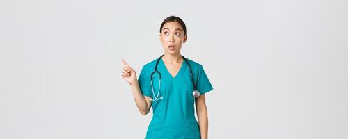 Covid-19, healthcare workers, pandemic concept. Surprised and amazed female asian physician looking and pointing upper left corner. Astonished korean doctor stare at banner with advertisement photo