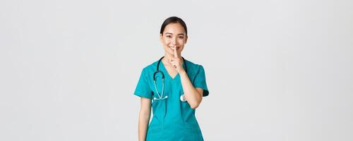 Covid-19, healthcare workers, pandemic concept. Cheerful, smiling female asian nurse in scrubs having secret, making surprise, showing shush gesture, shhh keep quiet, standing white background photo
