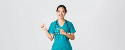 Covid-19, healthcare workers, pandemic concept. Intrigued beautiful asian female intern, nurse in scrubs looking and pointing upper left corner with curious dreamy smile, white background photo