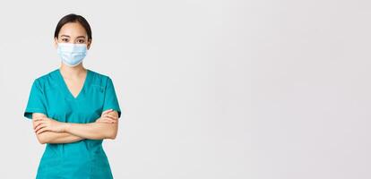 Covid-19, coronavirus disease, healthcare workers concept. Smiling confident asian female doctor, physician making checkup, wearing scrubs and medical mask, looking determined, white background photo