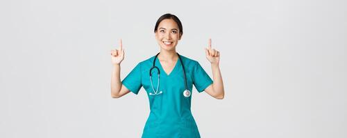 COVID-19, cuidado de la salud trabajadores, pandemia concepto. sonriente hermosa asiático hembra enfermero, médico en matorrales señalando y mirando arriba con satisfecho sonrisa, contento a espectáculo increíble promoción oferta, blanco antecedentes foto