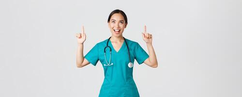COVID-19, cuidado de la salud trabajadores, pandemia concepto. divertido alegre asiático hembra enfermero, médico o cirujano en matorrales señalando dedos y mirando arriba con satisfecho rostro, en pie blanco antecedentes foto
