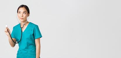 Covid-19, healthcare workers and preventing virus concept. Young focused asian female doctor, physician in scrubs, examine patient lungs, using stethoscope, standing thoughtful white background photo