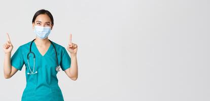 Covid-19, coronavirus disease, healthcare workers concept. Pleased cunning asian female nurse, doctor in medical mask and scrubs, looking and pointing fingers up, white background photo