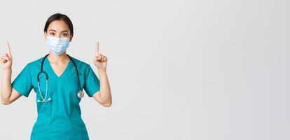 Covid-19, coronavirus disease, healthcare workers concept. Young professional asian female doctor, nurse in medical mask and scrubs, pointing fingers up, showing way, advertising photo
