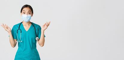 Covid-19, coronavirus disease, healthcare workers concept. Surprised and amazed asian female nurse, physician in medical mask and scrubs raising hands up excited, hear amazing news, white background photo