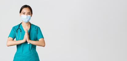 Covid-19, coronavirus disease, healthcare workers concept. Hopeful smiling asian female physician, doctor in medical mask and scrubs hold hands in praying gesture, thanking, white background photo