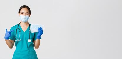 Covid-19, coronavirus disease, healthcare workers concept. Smiling asian female doctor, physician or nurse in rubber gloves and scrubs pointing finger at medical masks for patients safety photo