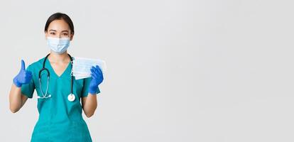 Covid-19, coronavirus disease, healthcare workers concept. Smiling asian doctor, nurse in scrubs and rubber gloves, showing thumbs-up and medical masks, white background photo