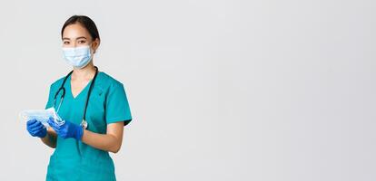 Covid-19, coronavirus disease, healthcare workers concept. Smiling friendly female nurse, physician in scrubs and rubber gloves provide patients with medical masks, white background photo