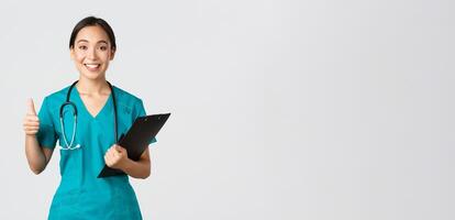 Healthcare workers, preventing virus, quarantine campaign concept. Smiling pretty asian doctor, female intern in scrubs show thumb-up, holding clipboard, bring positive results, white background photo