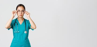 Covid-19, healthcare workers, pandemic concept. Confident serious-looking and determined asian female doctor, nurse in scrubs put on glasses, ready for shift in emergency room, white background photo