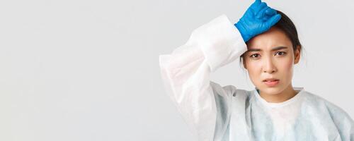 Covid-19, coronavirus disease, healthcare workers concept. Young tired asian female doctor take-off personal protective equipment and wipe sweat, looking exhausted after long shift in hospital photo