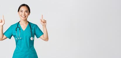 COVID-19, cuidado de la salud trabajadores, pandemia concepto. sonriente hermosa asiático hembra enfermero, médico en matorrales señalando y mirando arriba con satisfecho sonrisa, contento a espectáculo increíble promoción oferta, blanco antecedentes foto