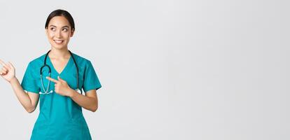 Covid-19, healthcare workers, pandemic concept. Intrigued beautiful asian female intern, nurse in scrubs looking and pointing upper left corner with curious dreamy smile, white background photo