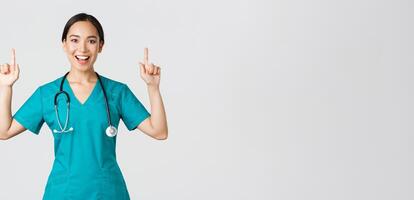 Covid-19, healthcare workers, pandemic concept. Smiling cheerful pretty nurse, female doctor or intern in scrubs pointing fingers up, showing banner, make announcement, demonstrate advertisement photo
