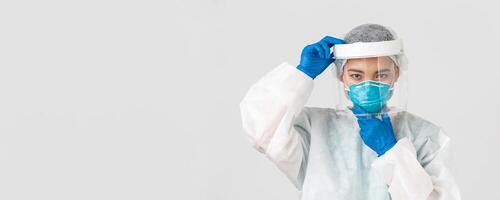 Covid-19, coronavirus disease, healthcare workers concept. Confident serious-looking female asian doctor, put on face shield and respirator, personal protective equipment, white background photo