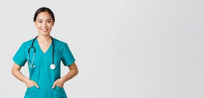 Covid-19, healthcare workers, pandemic concept. Confident smiling pretty asian female doctor, physician looking determined and upbeat, holding hands in pockets of scrubs, examine patients in clinic photo