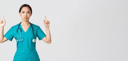 Covid-19, healthcare workers, pandemic concept. Gloomy disappointed asian female doctor, physician in scrubs pointing fingers up and grimacing displeased, standing white background photo