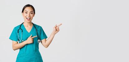 Covid-19, healthcare workers, pandemic concept. Glad smiling, beautiful asian doctor, female therapist in scrubs or nurse pointing fingers upper right corner, showing way to patient, white background photo