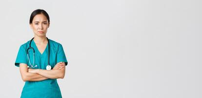 Covid-19, healthcare workers, pandemic concept. Skeptical and reluctant asian female doctor, tired nurse in scrubs cross arms and frowning, smirk displeased, standing white background photo